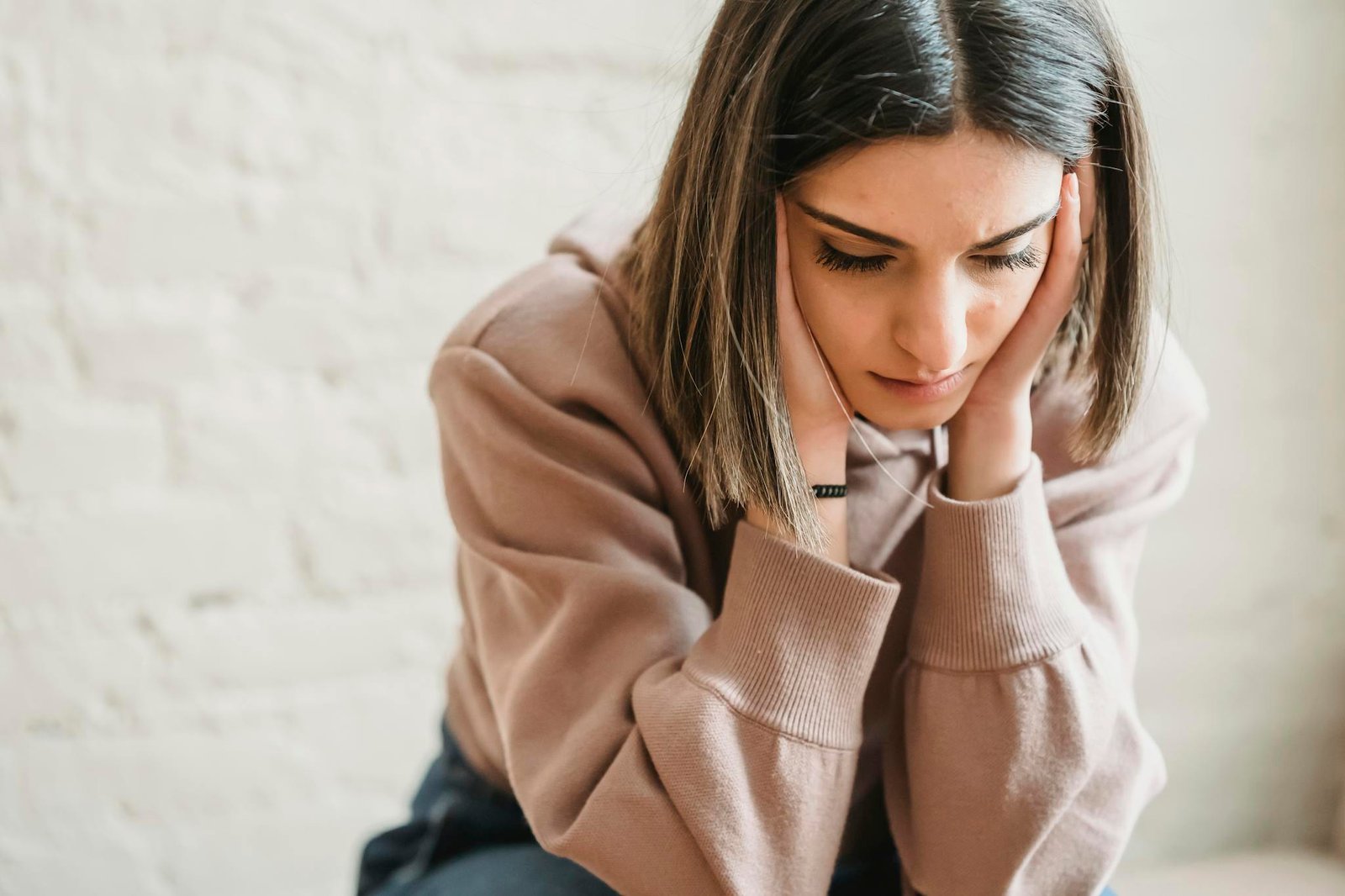 Crop unhappy female in casual wear touching cheeks and looking down while sitting near white wall in light room at home