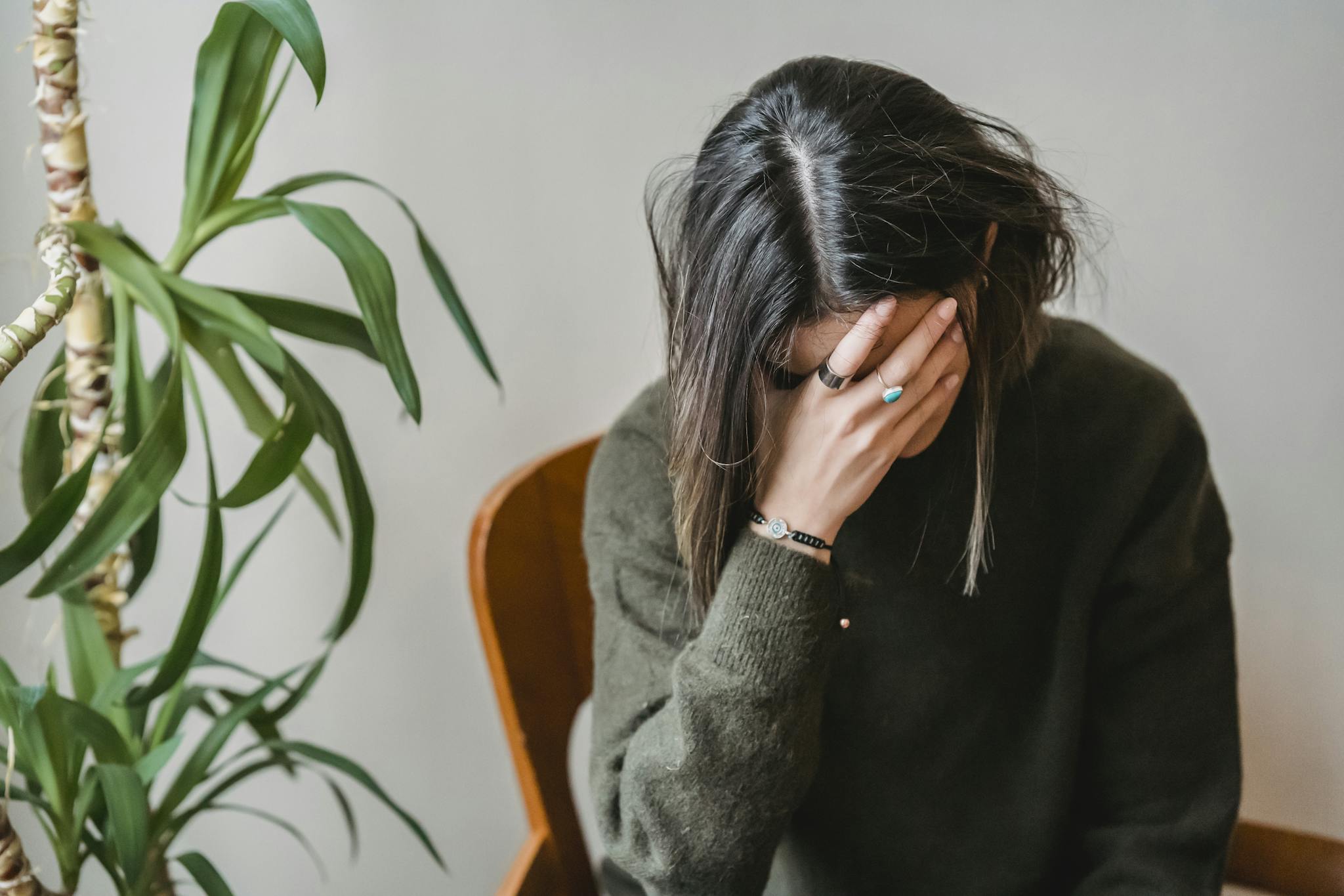 Unrecognizable young upset female millennial with dark hair in stylish sweater covering face with hand while siting on chair at home